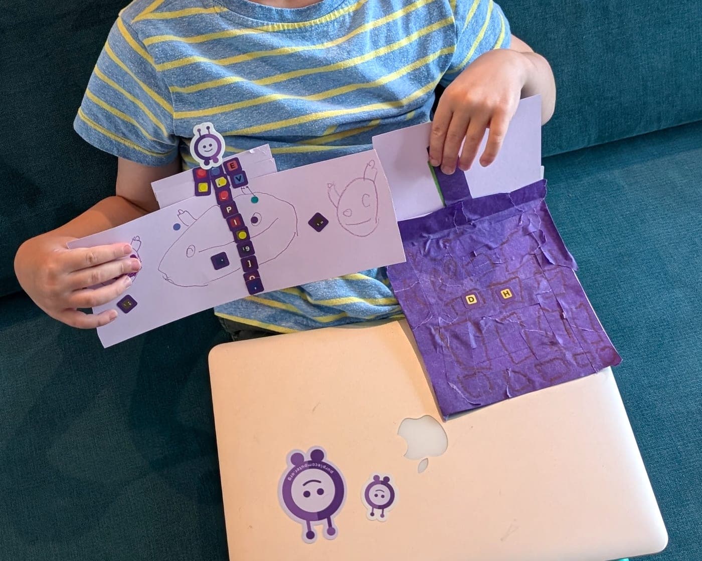 A kid holding up a Purple Computer made of paper: a tissue-paper laptop with letter stickers for keys, and a hand-drawn sign, sitting on a real laptop running Purple